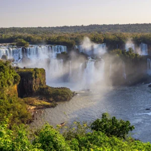 Vista panorâmica das Cataratas do Iguaçu sob a luz do pôr do sol, destacando as quedas d'água envoltas em névoa e a exuberante vegetação da Mata Atlântica no Parque Nacional.