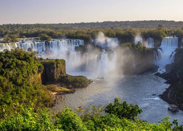 Vista panorâmica das Cataratas do Iguaçu sob a luz do pôr do sol, destacando as quedas d'água envoltas em névoa e a exuberante vegetação da Mata Atlântica no Parque Nacional.